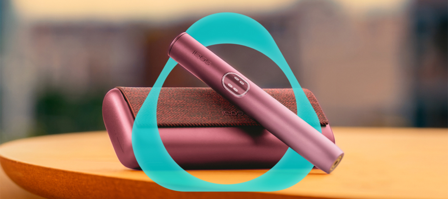 Dispositivo IQOS ILUMA i PRIME Vermelho Escuro sobre carregador de bolso, sobrepostos com logótipo IQOS, numa mesa, fundo com ambiente exterior desfocado